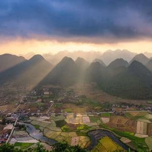 Rayos de sol atraviesan nubes oscuras iluminando un valle de montañas verdes, campos de arroz en terrazas y un pequeño pueblo.