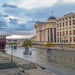 Una vista lungo il fiume di una città con una nave di legno attraccata accanto a un grande edificio neoclassico sotto un cielo nuvoloso.