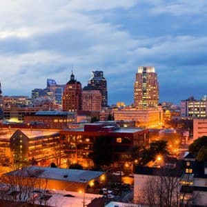 Un horizonte urbano con rascacielos y edificios iluminados resplandece con luces cálidas bajo un cielo crepuscular nublado.