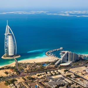 Veduta aerea di un hotel a forma di vela su una piccola isola artificiale, collegata da un ponte a una costa sabbiosa e a una città.