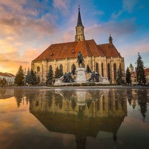 Una grande chiesa di pietra e un monumento riflessi in una grande pozzanghera in una piazza cittadina sotto un cielo al tramonto colorato.