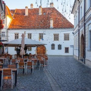 Un caffè all'aperto vuoto con tavoli e sedie di legno si trova su una strada acciottolata tra edifici storici all'alba.