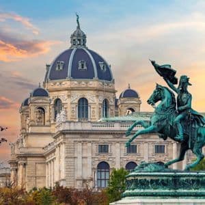 A bronze equestrian statue on a pedestal in front of a large, ornate building with a prominent dome under a sunset sky.