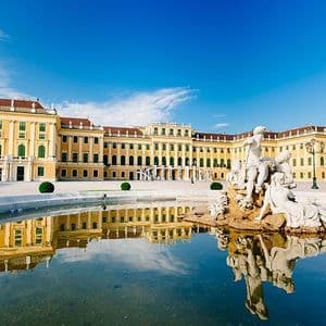 A large yellow palace with green shutters stands behind a fountain with white marble statues, reflected in the water on a clear day.