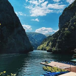 Kayak colorati si trovano su un molo di legno lungo un fiume che scorre attraverso un canyon ripido e alberato sotto un cielo azzurro e nuvoloso.