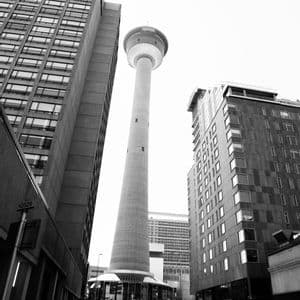 Une vue en noir et blanc, en contre-plongée, d'une haute tour d'observation encadrée par des gratte-ciel modernes dans une rue de ville.
