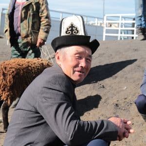 Un homme souriant coiffé d'un chapeau brodé traditionnel est assis en plein air avec un autre homme et un mouton brun en arrière-plan.