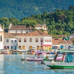 Colorful boats float in a bay in front of a coastal town with colonial-style buildings and a church, with a lush, green hill in the background.