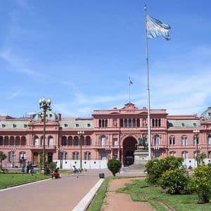 Un gran edificio gubernamental rosa se alza detrás de un parque, con una bandera argentina ondeando en un alto mástil bajo un cielo azul brillante.