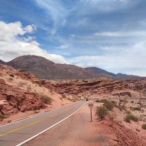 Una strada asfaltata con un cartello di limite di velocità si snoda attraverso un paesaggio desertico di formazioni rocciose rosse e montagne sotto un cielo blu e nuvoloso.