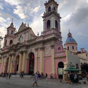 Una cattedrale rosa riccamente decorata, con colonne color crema e due campanili gemelli, si erge su una piazza acciottolata mentre le persone passeggiano.