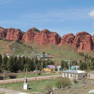Un petit village avec une mosquée est niché au pied de grandes formations rocheuses rouges superposées, sur une colline verdoyante sous un ciel bleu.