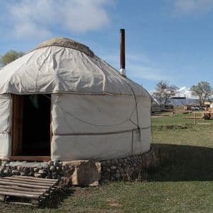 Une yourte blanche traditionnelle avec une base en pierre se trouve dans un camp herbeux, avec des montagnes enneigées visibles en arrière-plan.