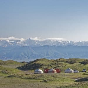 Un camp de yourtes avec des véhicules nichés dans des collines verdoyantes devant une chaîne de montagnes enneigée et lointaine.