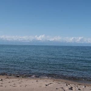 Une plage de sable rencontre l'eau bleue calme d'un lac, avec des montagnes enneigées visibles au loin sous un ciel clair.