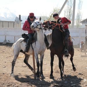 Deux hommes à cheval, l'un sur un cheval blanc et l'autre sur un cheval brun, se font face dans un enclos poussiéreux avec des montagnes en arrière-plan.
