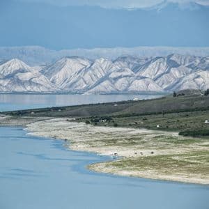 Un vaste lac bleu serpente le long d'une rive verdoyante, bordé de montagnes claires et striées, et de lointains sommets enneigés.