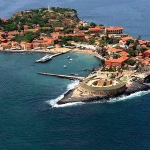 An aerial view of a small island with red-roofed buildings, a harbor with boats, and a stone fort in the blue sea.