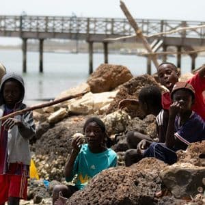 A group of children sitting and playing on a rocky shore, with a wooden pier spanning the water in the background.