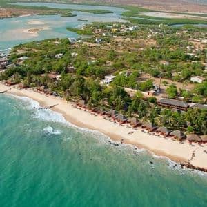 An aerial view of a coastal resort village with thatched-roof huts along a sandy beach and turquoise ocean.