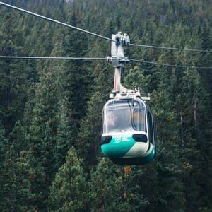 Une télécabine bleu sarcelle et blanche survole une dense forêt de pins recouvrant un flanc de montagne.