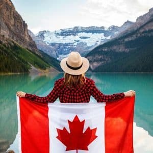Une personne de dos, vêtue d'une chemise à carreaux et d'un chapeau, tient un drapeau canadien tout en admirant un lac alpin turquoise et des montagnes.
