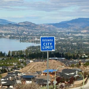 Un panneau bleu "Kelowna CITY LIMIT" sur une colline surplombe une ville, un lac et des montagnes sous un ciel nuageux.