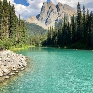 Un lac turquoise avec une rive rocheuse est entouré d'une forêt de pins, avec un sommet montagneux s'élevant à l'arrière-plan sous un ciel nuageux.
