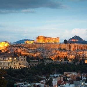 Una antigua ciudadela, brillantemente iluminada con luces doradas, se asienta sobre una colina rocosa con vistas a un paisaje urbano al anochecer.