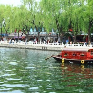 Un bateau rouge traditionnel, couvert, avec des passagers, vogue sur un canal le long d'une promenade arborée, animée par des personnes et des bâtiments.