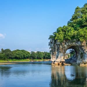 Une grande formation rocheuse couverte d'arbres avec une arche naturelle plongeant dans une large rivière sous un ciel bleu clair.