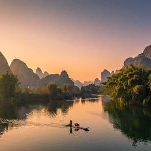 Une personne sur un radeau en bambou avance le long d'une rivière calme, entourée de montagnes karstiques et d'arbres luxuriants, au lever du soleil.