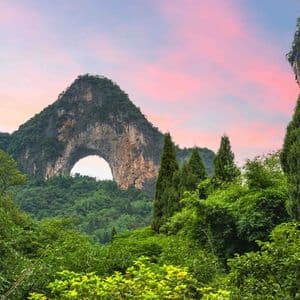 Une montagne avec une grande arche naturelle s'élève au-dessus d'une forêt verte dense sous un ciel rose et violet.