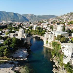 Una vista aerea di un vecchio ponte di pietra che attraversa un fiume turchese in una città storica, con case che si arrampicano sulle verdi colline sullo sfondo.