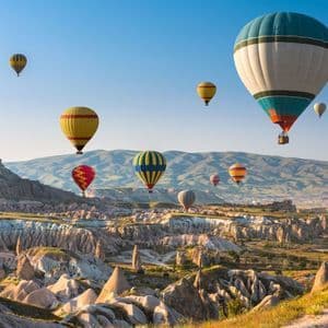 Mongolfiere colorate sorvolano un paesaggio di formazioni rocciose uniche e abitazioni rupestri durante un'alba dai colori pastello.