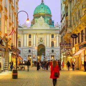 A busy cobblestone street at dusk, with people walking past illuminated shops towards a grand building with a green dome.
