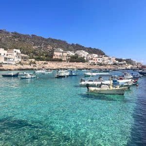 Small boats are moored in a calm bay with clear turquoise water, with a hillside town visible in the background under a blue sky.