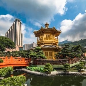 Une pagode dorée traditionnelle et un pont orange dans un bassin de jardin, avec une silhouette de ville moderne en arrière-plan sous un ciel bleu.