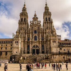 La ornamentada fachada de una gran catedral de piedra se eleva sobre una animada plaza llena de gente bajo un cielo parcialmente nublado.