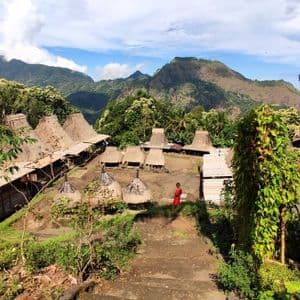 Ein traditionelles Dorf mit Reetdachhütten in einem grünen Bergtal, mit einer Person in Rot, die auf einem Pfad steht.