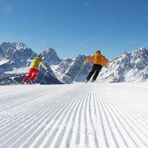 Two skiers descend a freshly groomed snow slope with a panoramic view of a snowy mountain range under a clear sky.