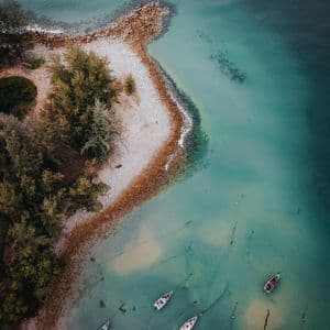 Luftaufnahme von oben: Eine geschwungene Küstenlinie mit einem Kiesstrand, Bäumen und mehreren kleinen Booten, die auf klarem, türkisfarbenem Wasser schwimmen.