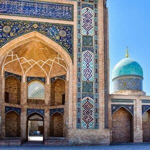 La ornamentada fachada de ladrillo de un edificio con intrincados mosaicos azules, amarillos y blancos y una cúpula turquesa bajo un cielo azul claro.