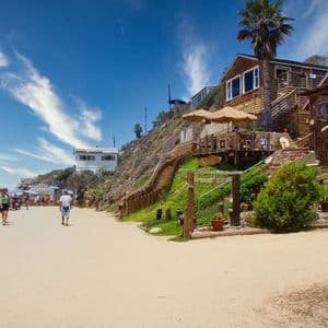 Le persone camminano lungo un sentiero sabbioso accanto a case sulla spiaggia in legno costruite su una collina sotto un cielo soleggiato e azzurro.