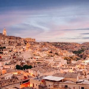Un borgo di antichi edifici in pietra e un campanile incastonato nella collina, illuminato al crepuscolo accanto a un burrone.