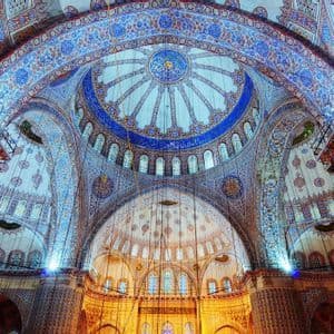 El interior de una mezquita con un techo abovedado intrincadamente decorado, con patrones azules y luces colgantes.