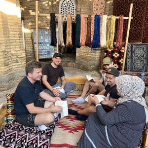 Un groupe WeRoad s'assied auprès d'une femme locale sur un banc, découvrant un savoir-faire artisanal dans un atelier aux fils colorés.