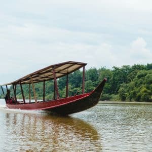 Una lancha de madera roja cubierta navega a toda velocidad por un río, con una densa orilla de jungla bajo un cielo nublado.