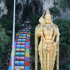 Una estatua gigante dorada de una deidad se alza junto a una empinada escalera de colores arcoíris que asciende por un acantilado rocoso.