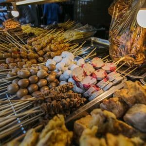 Variedad de brochetas de comida callejera, incluyendo huevos de codorniz, albóndigas y pasteles de pescado, exhibidas en bandejas de metal en un puesto de mercado.